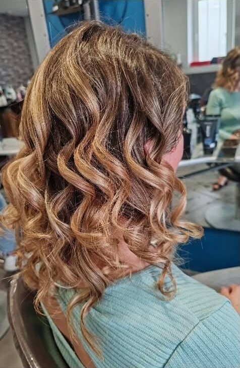 Cheveux mi-longs bouclés aux reflets blonds et dorés, vus de dos, dans un salon de coiffure aux murs bleus.
