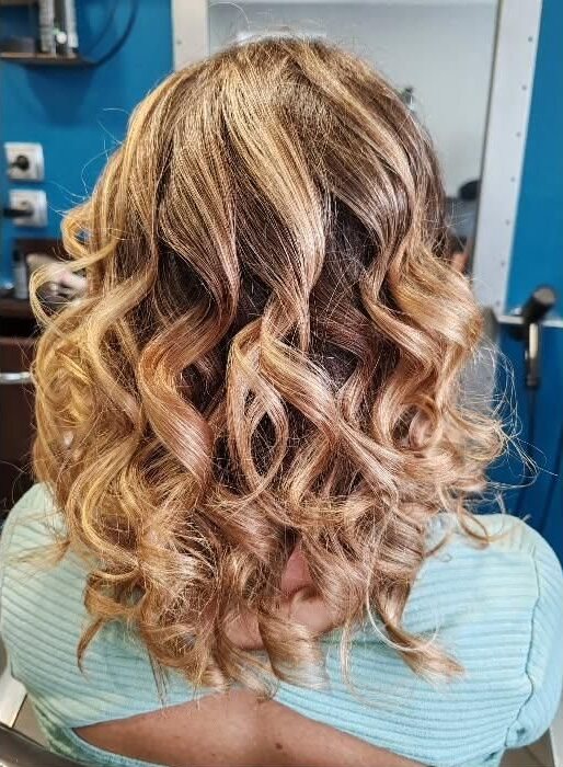 Cheveux mi-longs bouclés aux reflets blonds et dorés, vus de trois quarts dos, dans un salon de coiffure.