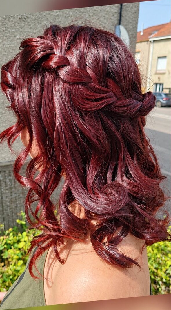 Vue de dos de cheveux mi-longs bouclés rouge bordeaux avec une tresse sur le côté, prise dehors.