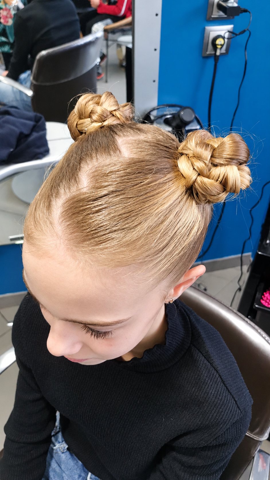 Une jeune fille assise dans un salon de coiffure porte deux chignons tressés sur le haut de la tête, vue de dessus.