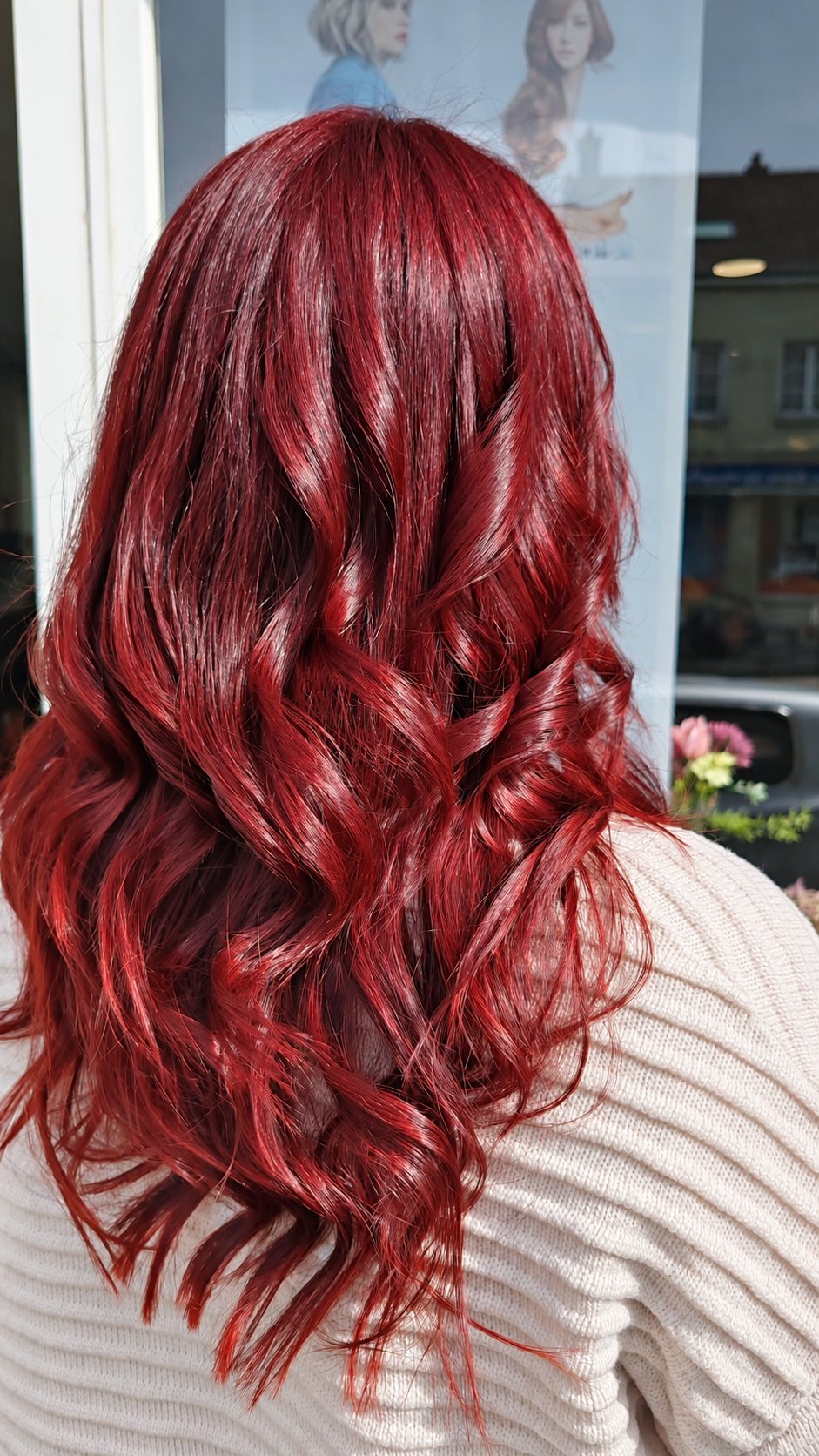 Vue de dos d'une personne aux longs cheveux ondulés d'un rouge intense et brillant, portant un pull beige, devant une vitrine de salon.