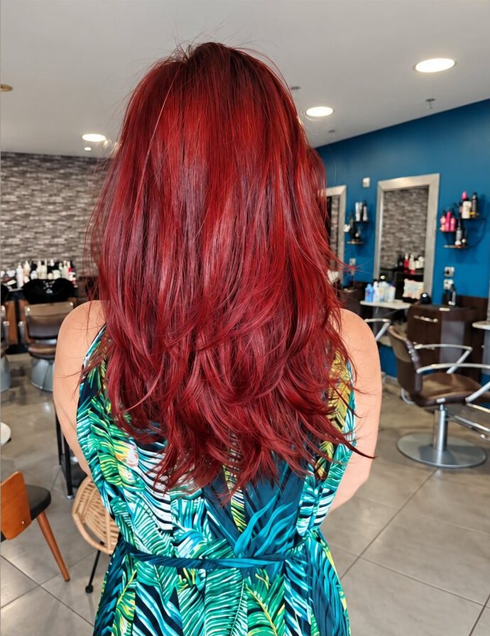 Vue de dos d'une femme en robe à motifs de feuilles tropicales, avec de longs cheveux dégradés d'un rouge intense, dans un salon de coiffure.