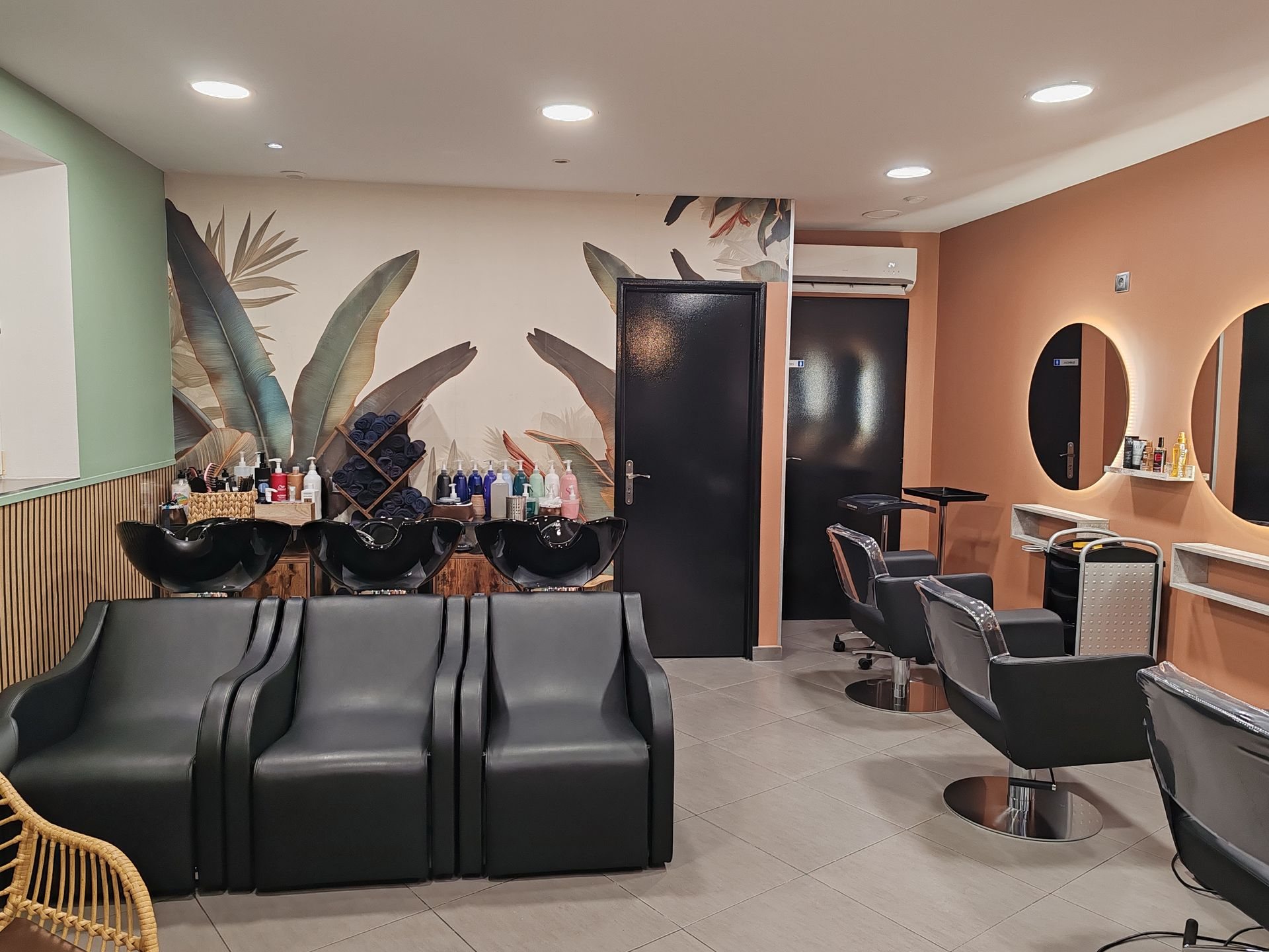 Intérieur d'un salon de coiffure aux murs vert et terracotta, avec trois bacs de lavage, des fauteuils noirs et des miroirs ronds lumineux.